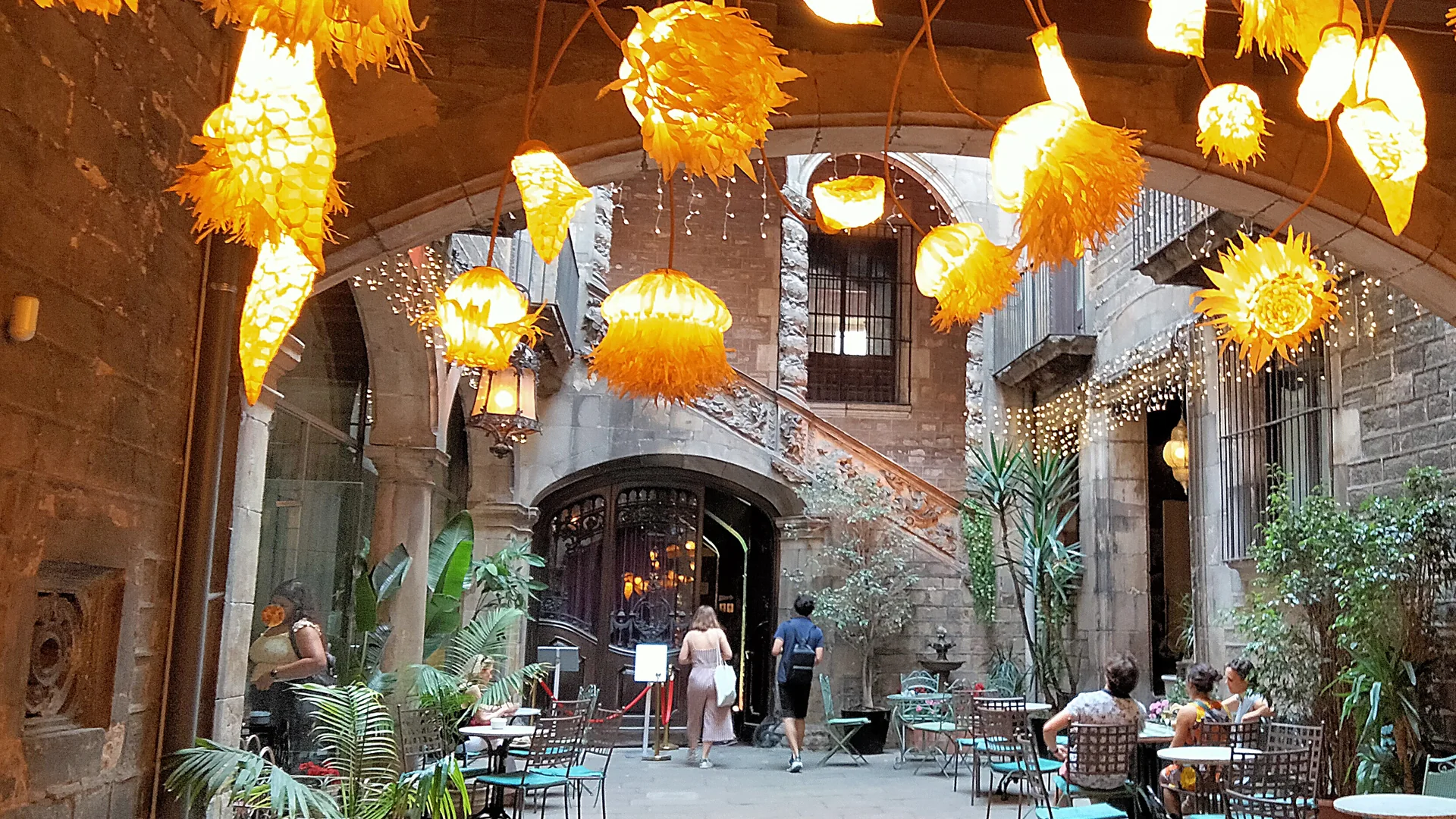 The courtyard of Palau Dalmases seen through a stone arch, with clusters of glowing yellow lanterns overhead, café tables below and the carved baroque staircase rising behind