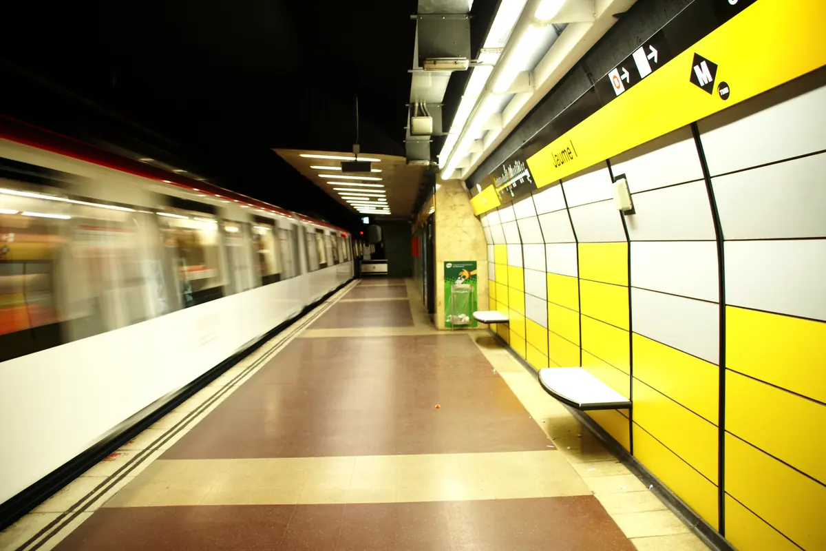 A yellow-tiled platform at Jaume I metro station in Barcelona with a train passing at speed