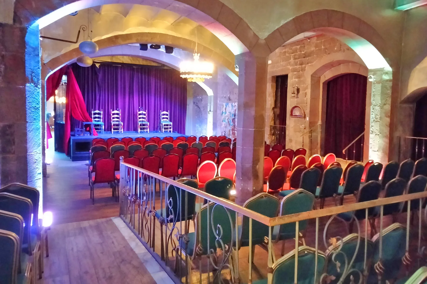 A vaulted hall in the palace with a stage, a purple curtain, rows of red chairs and a wrought-iron railing